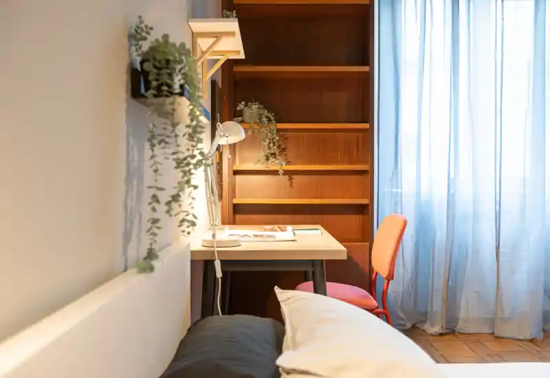 Bright, cozy bedroom corner with a visible bed foreground, a small desk and shelf with plants beside a sheer curtained window — ideal for a compact, well-styled rental.