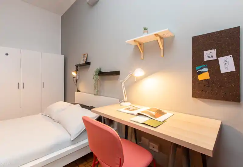 Bright, modern bedroom with a single bed, a wooden desk and a warm desk lamp — ideal for students or professionals seeking a compact, cozy workspace and sleeping area.