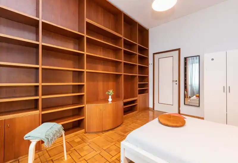 Bright, neat bedroom with a large built-in wooden shelving unit, a white bed with a decorative round cushion and parquet flooring.
