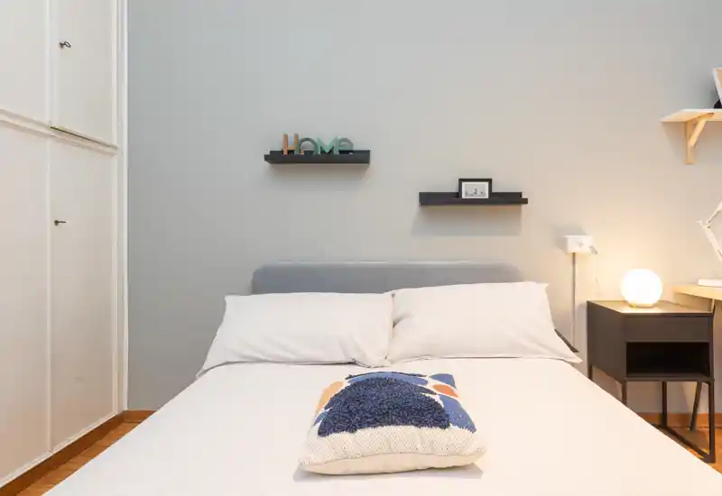 Camera da letto moderna e luminosa con letto matrimoniale centrato, biancheria chiara e un cuscino decorativo, comodino con lampada e mensole minimal.