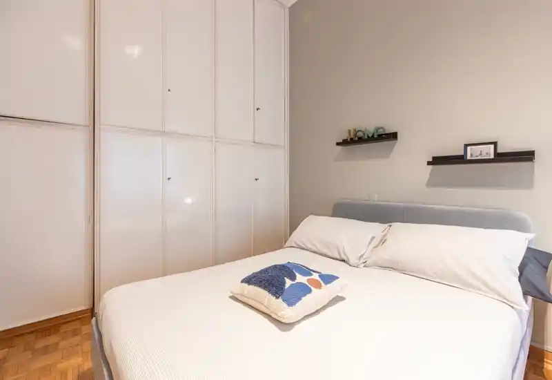 Camera da letto luminosa e ordinata con letto completo, testiera grigia e armadi a muro; un cuscino decorativo e mensole a parete creano un’atmosfera tranquilla.