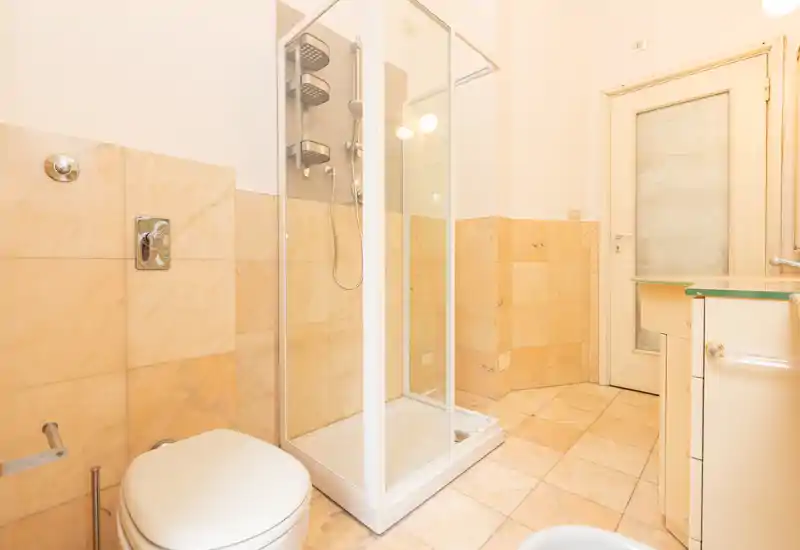 Clean, bright bathroom with a corner shower cabin, toilet, and sink; warm beige tiles and good natural/artificial lighting create a welcoming, functional space.