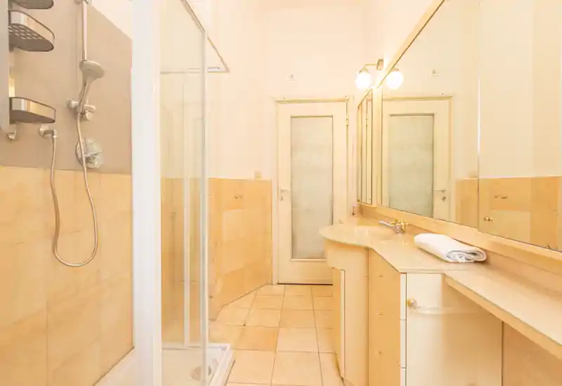 Bright, well-lit bathroom with a corner shower cabin, tiled floor and walls, and a long vanity with mirror and integrated sink.