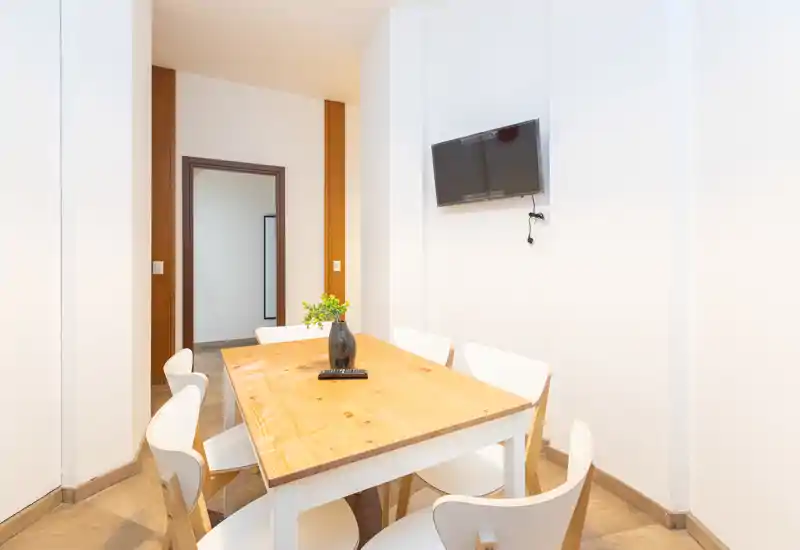 Bright, compact dining area with a wooden table, six modern white chairs and a wall-mounted TV — ideal for casual meals in a minimalist apartment.