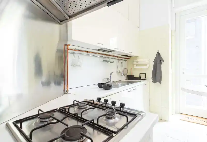 Bright modern kitchen with a four-burner gas stove, stainless steel backsplash, white cabinetry and a sink area; natural light spills in from a door/window, creating a clean, airy cooking space.