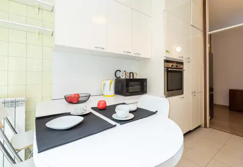 Bright modern kitchen corner with a small round dining table, built-in oven, microwave and glossy white cabinets; simple table setting and decorative fruit provide a welcoming touch.