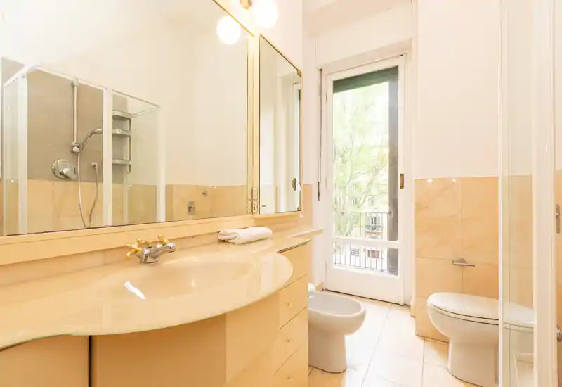 Bright, well-maintained bathroom with a large mirror, sink with countertop, shower enclosure, bidet and toilet. Natural light enters through a glass door leading to a small balcony or exterior view.