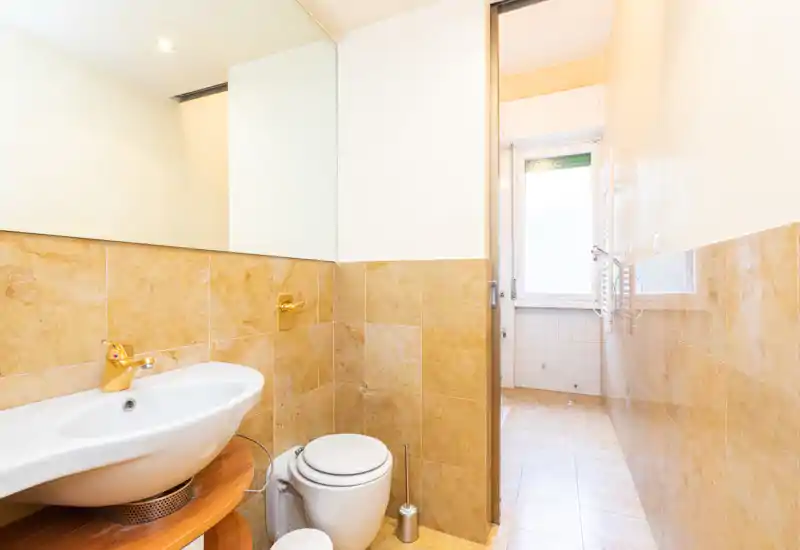 Small bathroom with beige tiled walls, modern sink with gold faucet, toilet and a large mirror; bright natural light from an adjacent window.