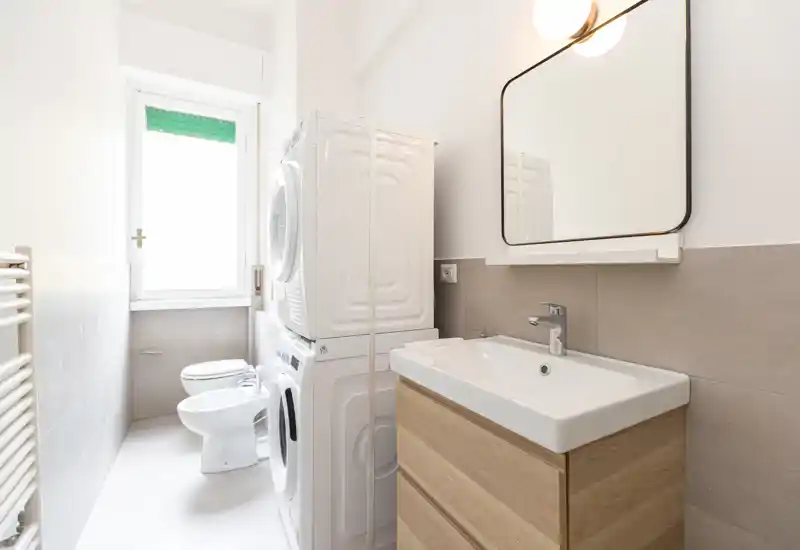 Bright modern bathroom with a wall-mounted mirror, sink with wood-effect vanity and stacked washer-dryer. Compact, practical layout with a bidet and toilet near a large window providing natural light.