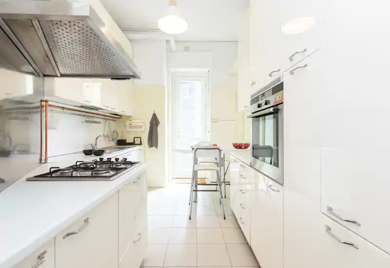 Bright modern galley kitchen with white cabinets, built-in oven, gas hob and a small breakfast table near a glass door letting in natural light.