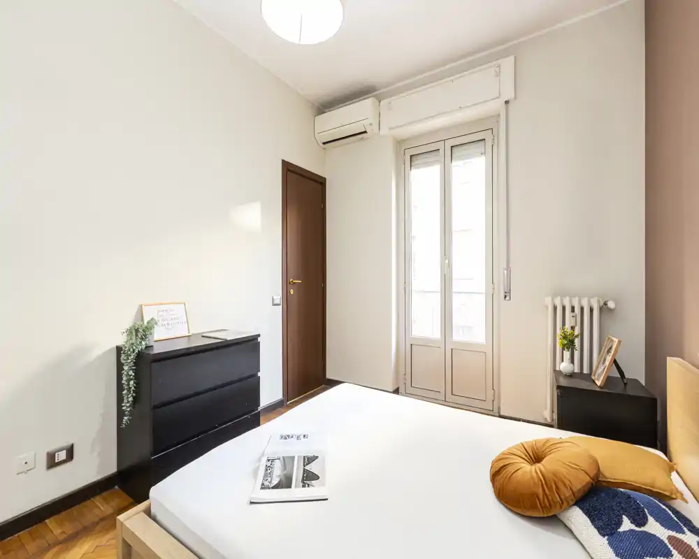 Bright, simply styled bedroom with a double bed in the foreground, a dresser and a balcony door letting in natural light. Neutral walls and minimal decor create a calm, inviting atmosphere ideal for rental listings.