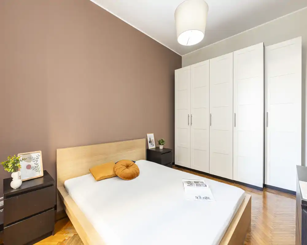 Bright, modern bedroom with a wooden bedframe, white linens, decorative cushions and a large white wardrobe. The warm accent wall and bedside plants give the room a cozy, stylish feel.