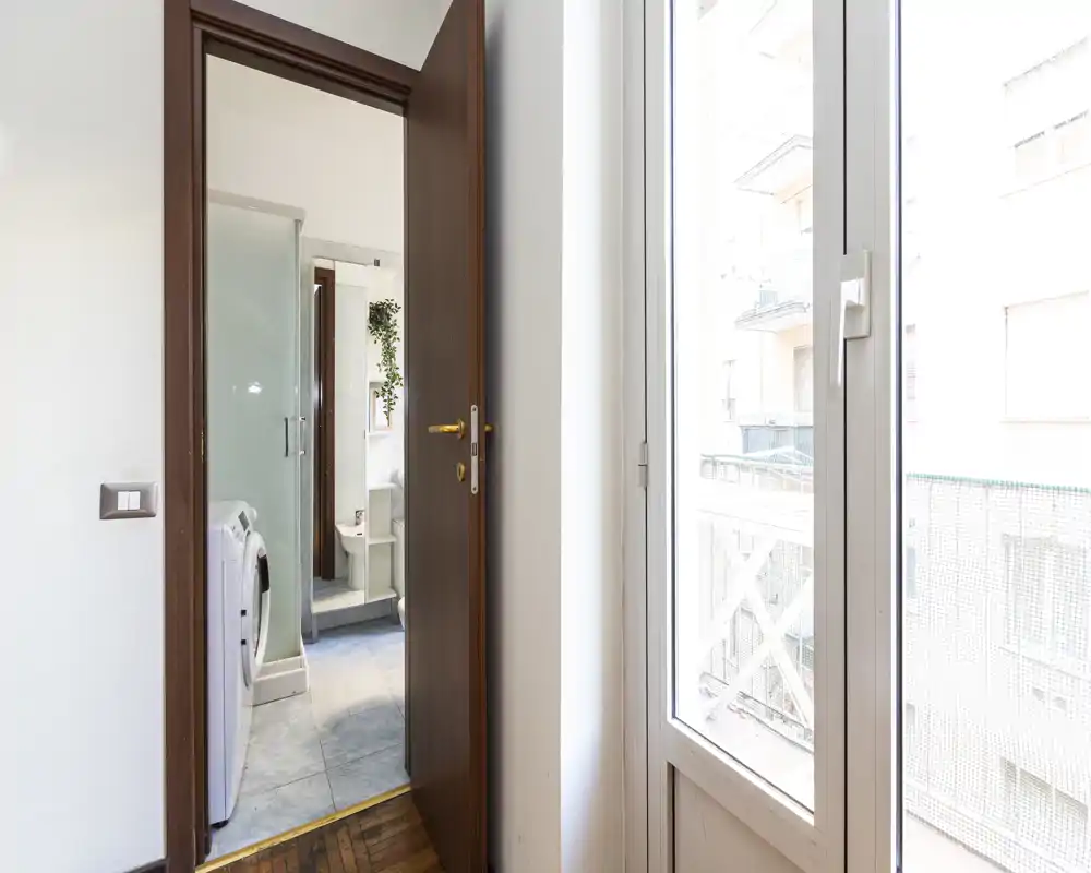 Small bathroom seen through an open door with a washing machine, shower enclosure and sink; bright natural light from a balcony window.