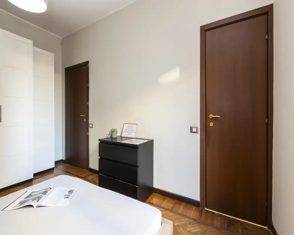Bright, minimalist bedroom with a visible mattress in the foreground, a black three-drawer chest, white wardrobe and wooden doors — clean lines and warm parquet flooring.