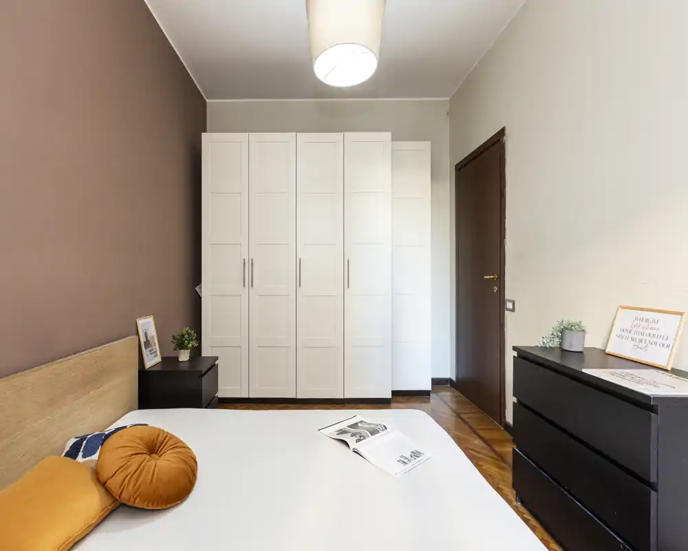 Bright, modern bedroom with a large bed in the foreground, white wardrobe against the wall and dark wood accents. Soft textiles and warm wall color create a cozy, well-styled atmosphere ideal for rental photos.