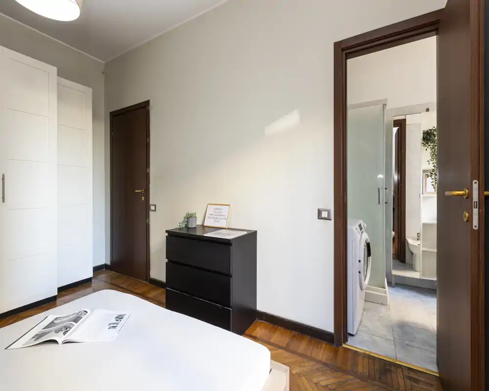 Spacious bedroom with a visible double bed, a black three-drawer dresser, wardrobe and direct access to an en-suite bathroom with laundry machine. Warm wooden flooring and neutral walls create a calm, well-lit sleeping area ideal for rental listings.
