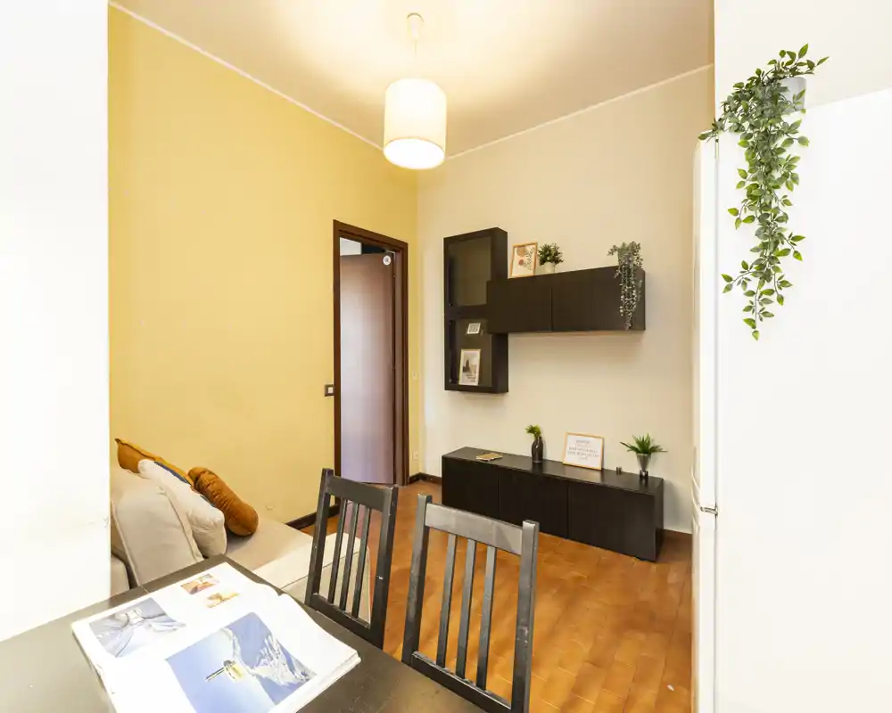 Well-lit living area with a small sofa, dining chairs in foreground and modern wall-mounted storage; warm tones and decorative plants create a comfortable atmosphere.