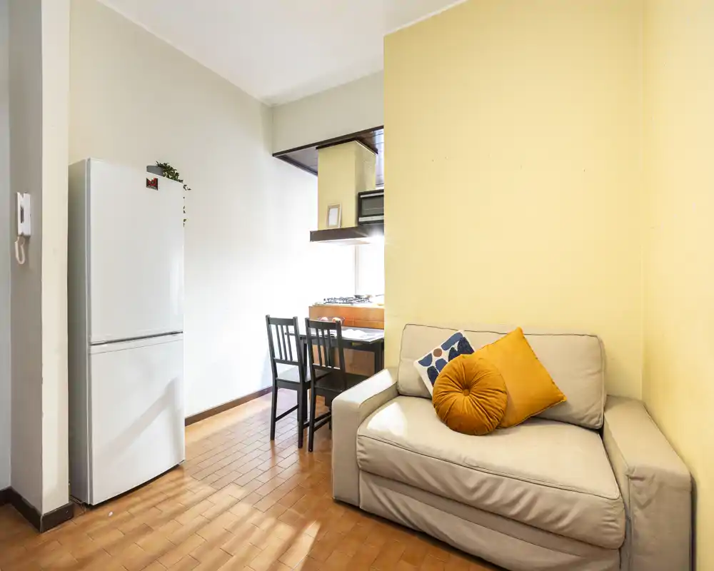Compact living corner with a comfortable cream sofa, colorful cushions and a small dining table by the kitchen; bright natural light and warm terracotta flooring create a cozy, functional space.