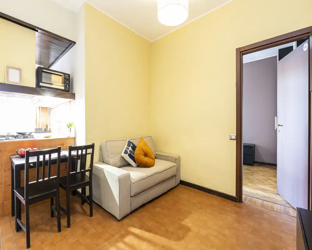 Small living area with a two-seater sofa, decorative cushions and a compact dining counter adjacent to the kitchenette. Bright, simply furnished space ideal for a studio or one-bedroom flat.
