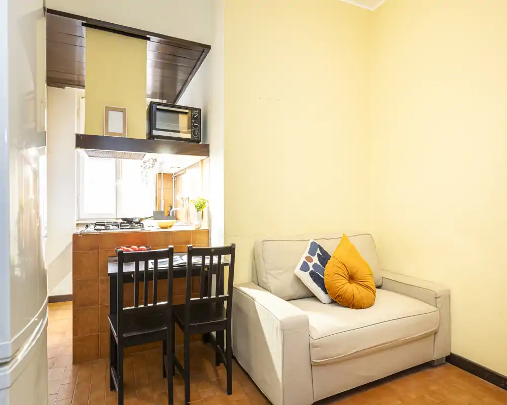Compact living area with a small sofa and decorative cushions beside an open kitchenette and a breakfast bar with two chairs — bright, tidy and ideal for a couple or single occupant.
