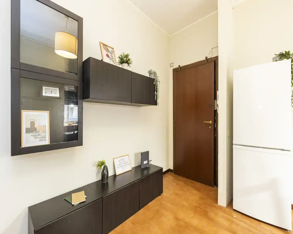 Well-lit entrance hallway with terracotta tiles, dark storage cabinets mounted on the wall and a white refrigerator near the door — tidy and practical entry area.