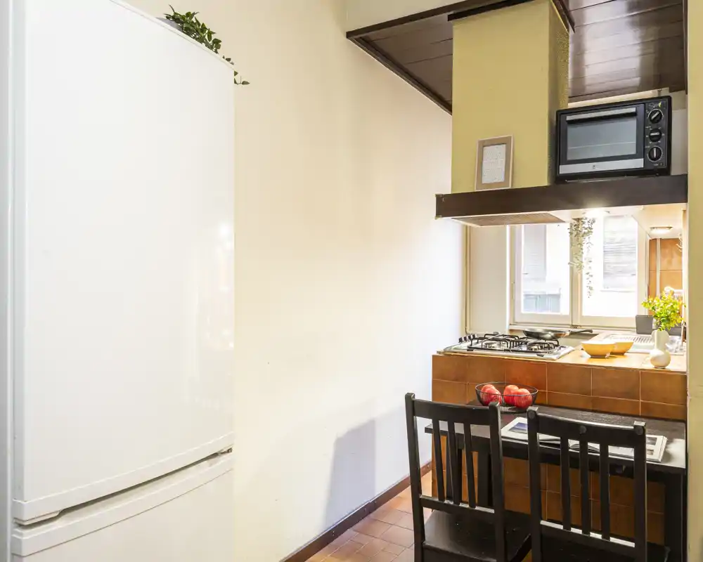 Compact, well-lit kitchen with a countertop dining spot, gas hob, microwave and fridge. Warm tiles and small decorative touches create a welcoming atmosphere.