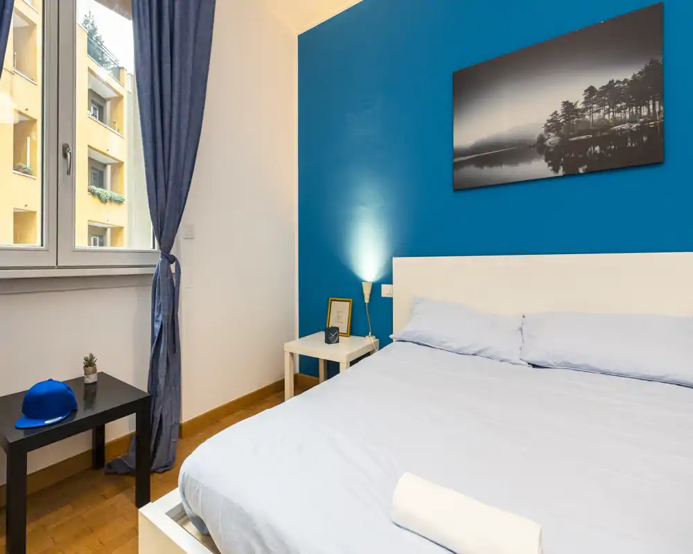 Camera da letto moderna e luminosa con parete blu, letto matrimoniale, comodino con lampada e cornice; luce naturale da una grande finestra.