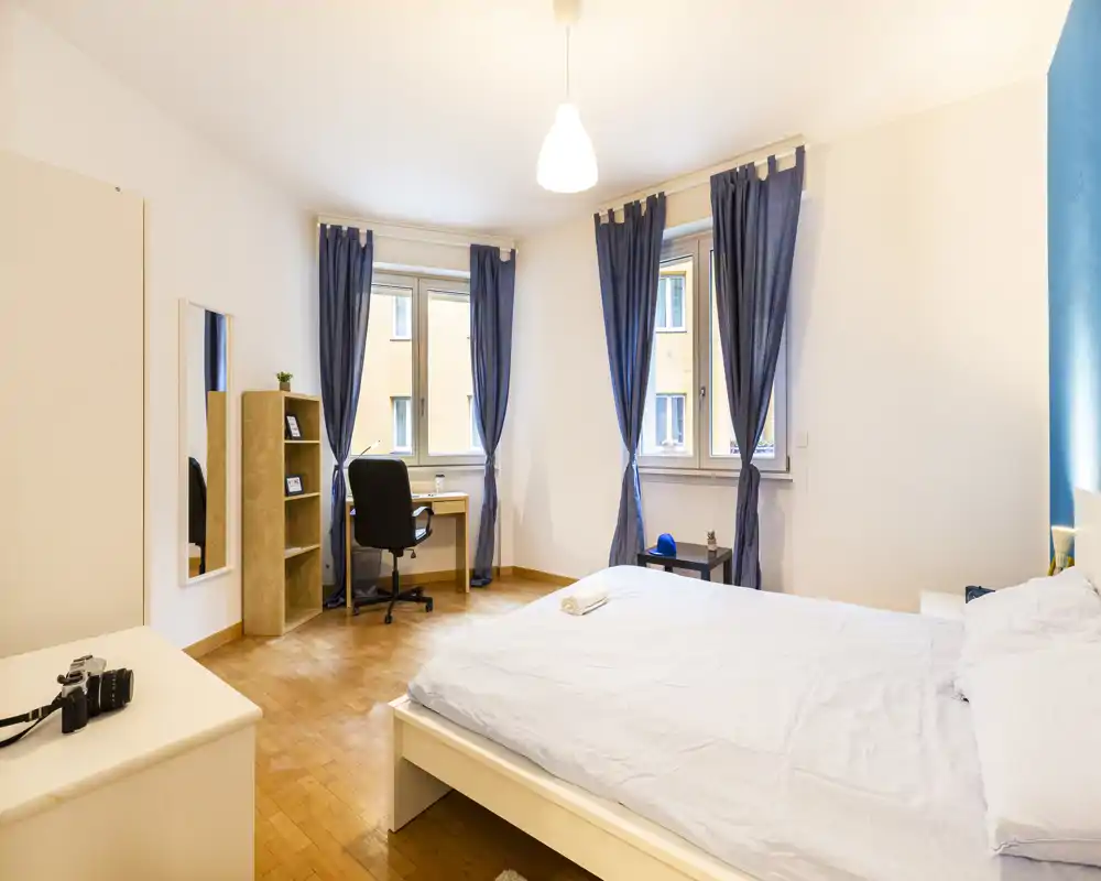 Ampia e luminosa camera da letto con letto matrimoniale, pavimento in parquet, scrivania vicino alla finestra e tende blu che creano un'atmosfera rilassante.