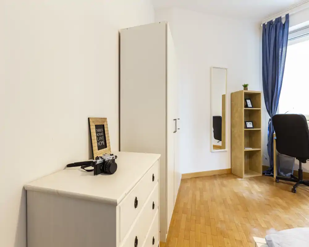 Angolo di camera da letto luminoso e ordinato con pavimento in legno chiaro, un comò, un armadio, una libreria e una scrivania vicino alla finestra. La luce naturale e l’arredamento essenziale lo rendono adatto a soggiorni brevi o lunghi.