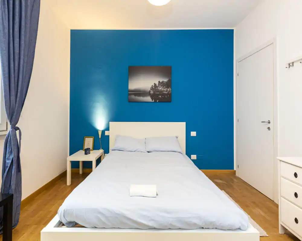 Camera da letto luminosa e moderna con letto matrimoniale al centro su una parete blu acceso, comodino e biancheria morbida — perfetta per annunci.
