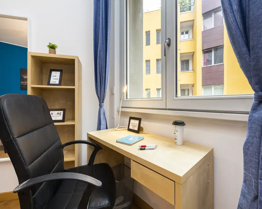 Angolo studio ordinato nella camera con scrivania in legno sotto una finestra ampia, poltrona da ufficio nera e mensole con cornici decorative — luce naturale e tende blu creano un'atmosfera rilassante.