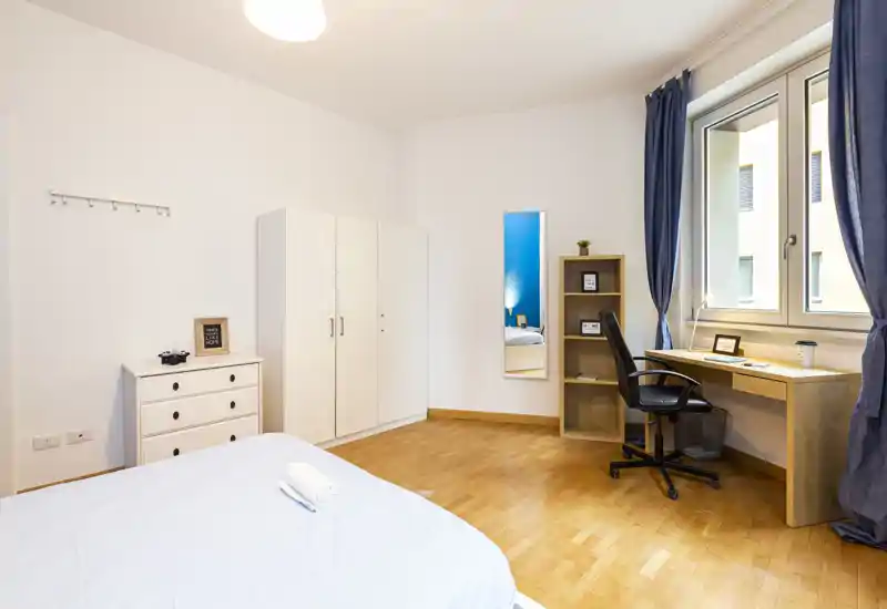 Camera da letto luminosa e spaziosa con letto grande, pavimento in legno e scrivania vicino alla finestra. Tende blu e arredi moderni e minimal creano un ambiente tranquillo e accogliente, perfetto per affitti brevi.