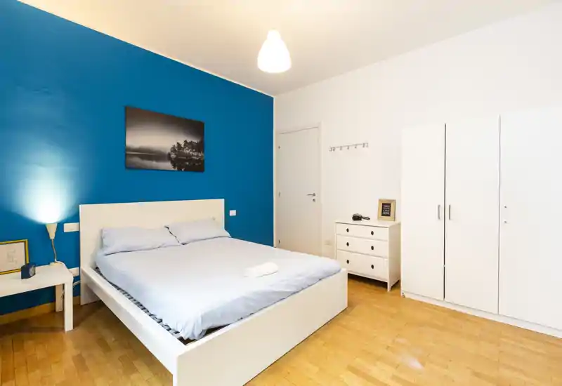 Camera da letto luminosa e moderna con un ampio letto matrimoniale, parete blu a contrasto e mobili bianchi. Spazio ordinato, ben illuminato e dallo stile semplice, ideale per annunci.