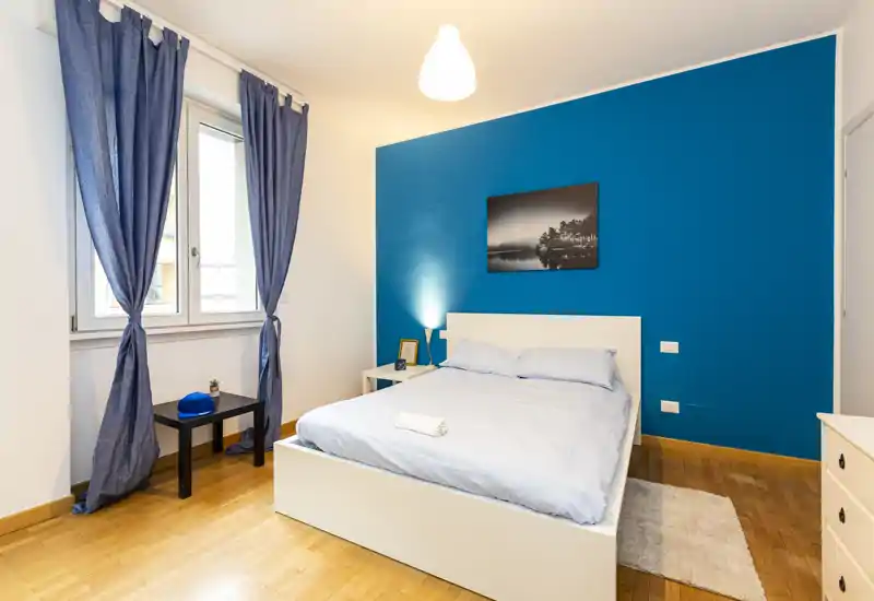 Camera da letto luminosa e moderna con letto matrimoniale, parete accentuata blu e luce naturale dalla finestra; letto ben rifatto, comodini e tessuti morbidi per un ambiente accogliente.