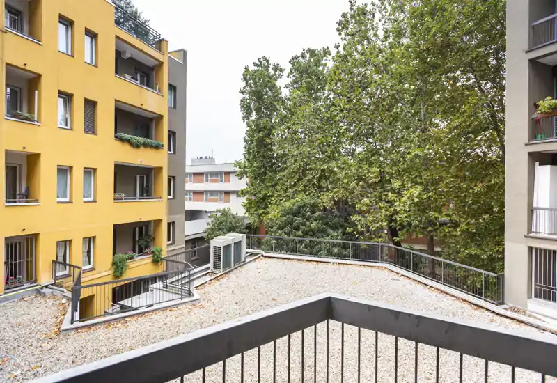 Balcone/terrazza con vista su un tetto in ghiaia delimitato da ringhiere, facciate di appartamenti vicini e alberi maturi che offrono privacy e verde.