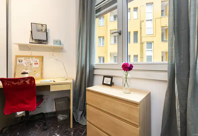 Zona giorno luminosa e ordinata con un piccolo angolo studio: scrivania in legno e sedia rossa accanto a un comò sotto una grande finestra; dettagli decorativi includono cornici e fiori freschi.