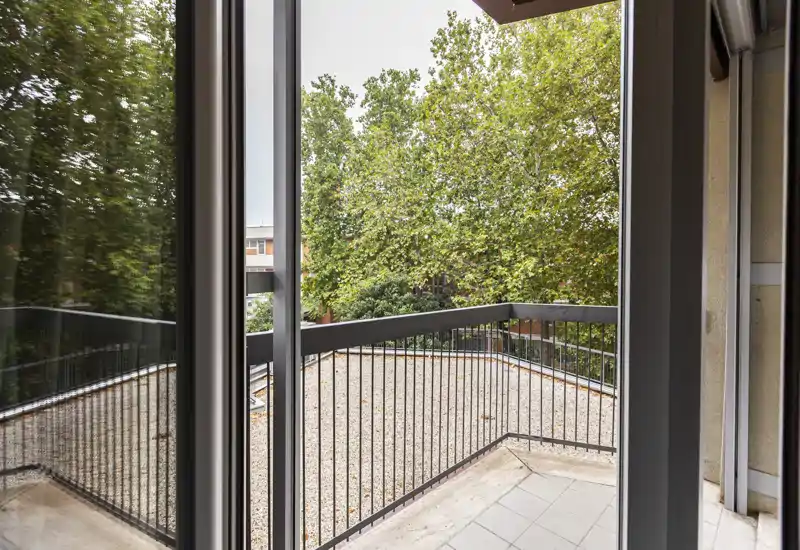 Balcone soleggiato inquadrato da porta scorrevole in vetro, con ringhiera in metallo e alberi sullo sfondo — perfetto per il caffè mattutino.