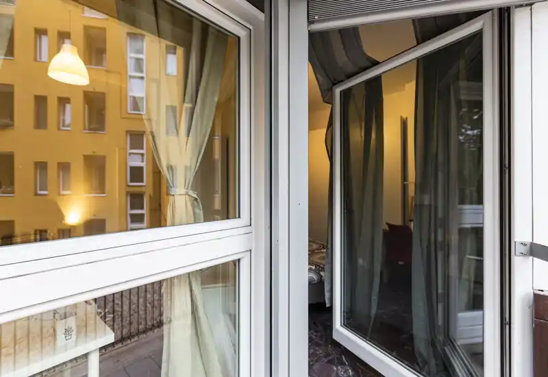 Vista attraverso la finestra/porta sul balcone verso una camera da letto con porta in vetro aperta, tende e luce calda interna. Il letto è parzialmente visibile e si notano tessili interni e riflessi esterni.