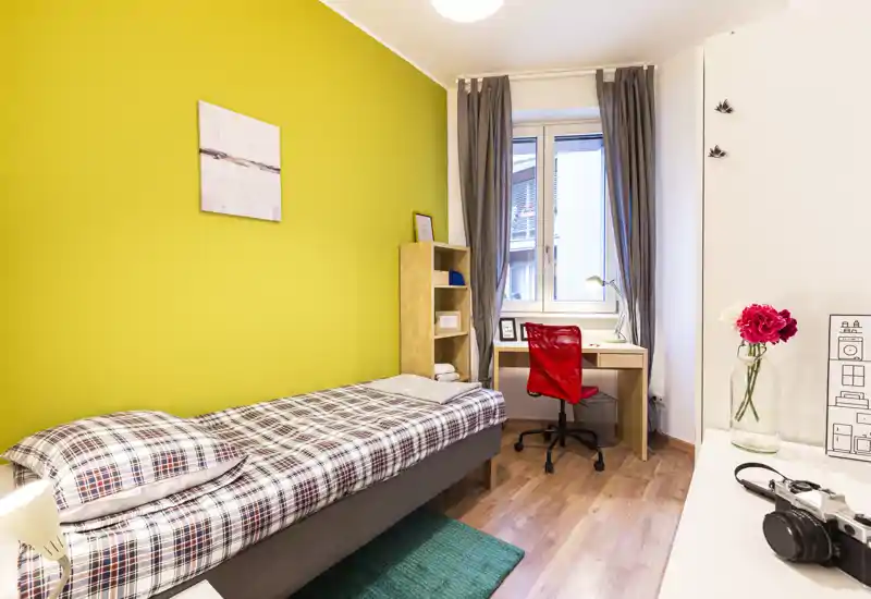 Chambre simple lumineuse et bien décorée avec lit à carreaux, bureau près de la fenêtre et sol en bois — idéale pour un étudiant.