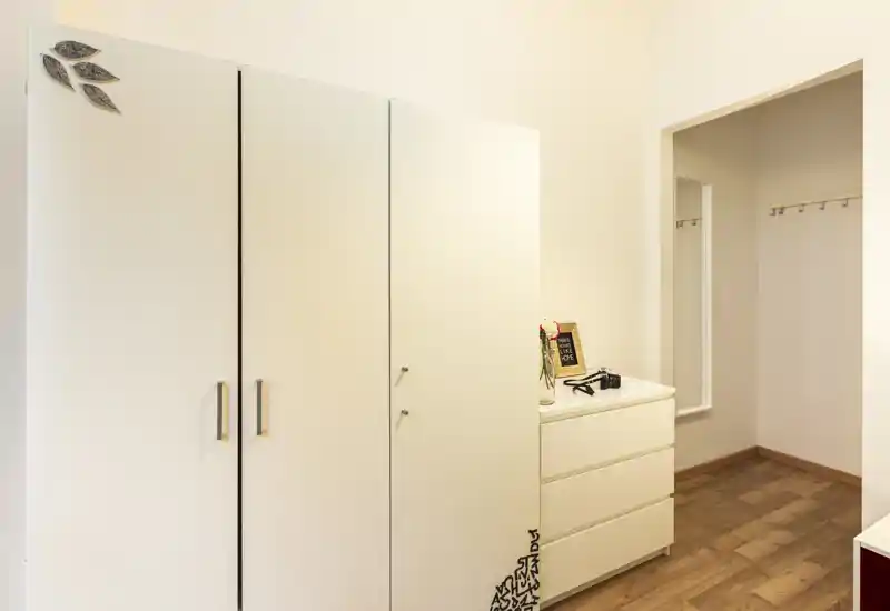 Ingresso ordinato con armadi bianchi, un comò e attaccapanni a parete; pavimento in legno caldo e pareti neutre creano uno spazio minimo e funzionale per riporre giacche e scarpe.