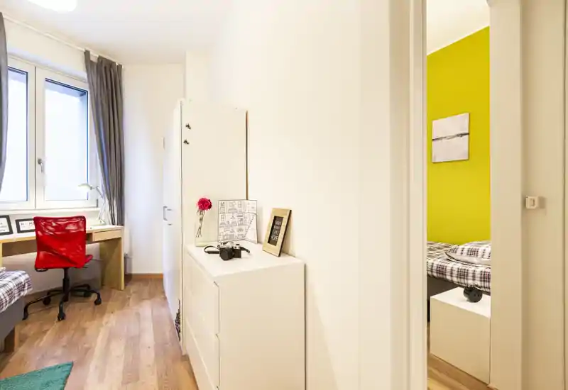 Coin de chambre bien éclairé visible à travers une porte, avec literie à carreaux, table de chevet et un espace bureau soigné au premier plan. Style moderne et lumineux, idéal pour une annonce locative.