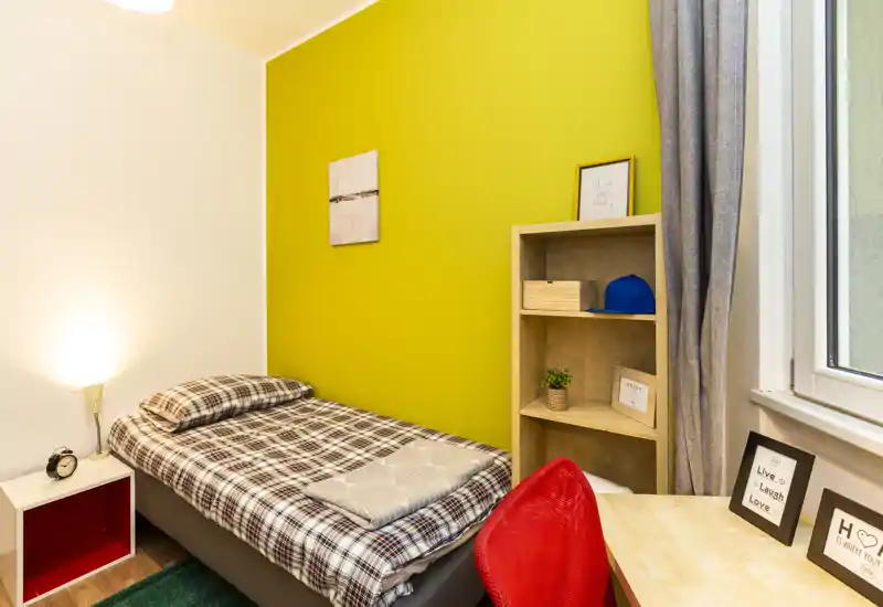 Chambre individuelle lumineuse et bien rangée avec lit à carreaux, lampe de chevet et petit bureau près de la fenêtre. Mur jaune vif en accent et étagères pratiques — idéale pour un étudiant ou une personne seule.