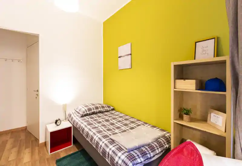 Chambre simple avec lit bien fait contre un mur jaune vif, table de chevet avec lampe et étagère offrant rangement et décorations — lumineuse, chaleureuse et bien rangée.