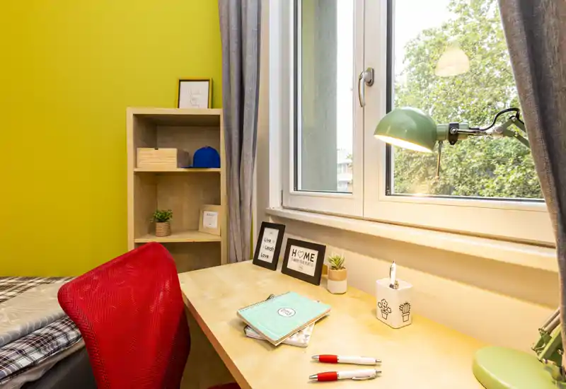 Coin chambre lumineux et soigné avec un bureau près de la fenêtre. On voit un bureau avec lampe et petits objets décoratifs, une chaise rouge et le bord du lit avec une couette à motifs.