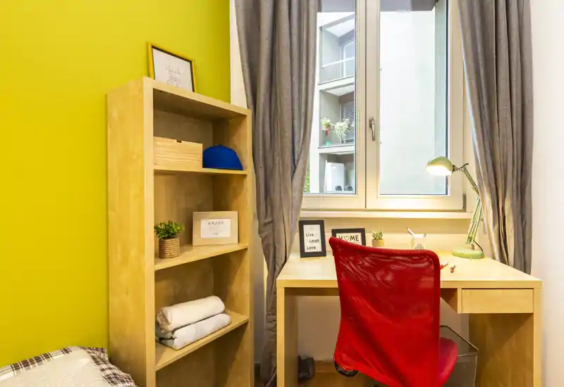 Angolo di camera da letto compatto con scrivania in legno vicino a una finestra luminosa, sedia rossa in mesh e scaffale aperto; piccole piante e teli piegati creano un ambiente ordinato e funzionale.