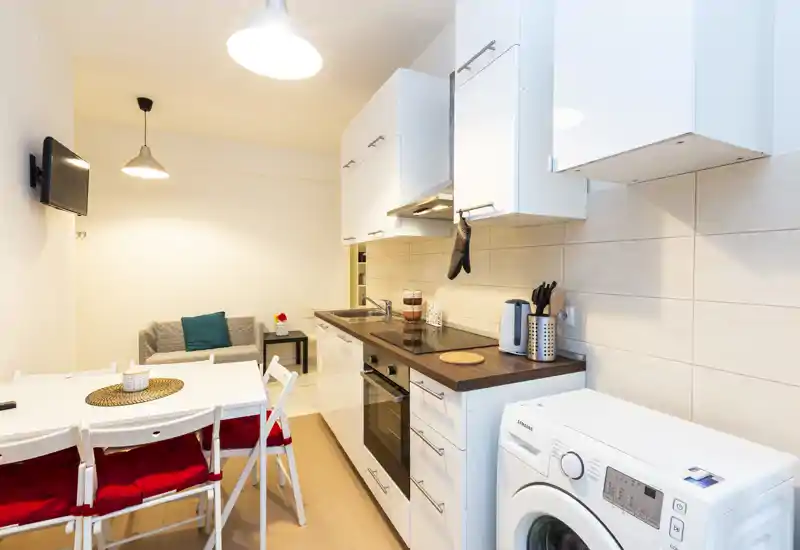 Zona giorno open-space con angolo cottura compatto. La stanza ha pensili bianchi, piano scuro in legno, lavatrice, un piccolo tavolo da pranzo con sedie con cuscini rossi e un divano due posti: luminosa e ordinata, ideale per affitti brevi.