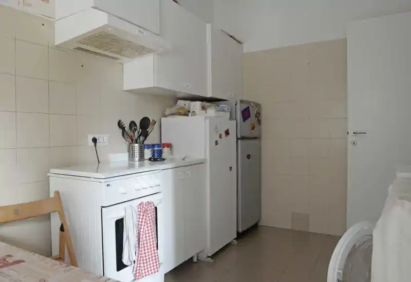 Angolo cucina compatto e ordinato con forno a libera installazione, pensili, frigorifero e utensili da cucina; piastrelle neutre e luce naturale creano uno spazio pratico e ben tenuto.