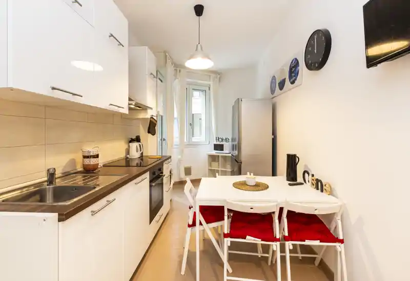 Cucina moderna e luminosa con mobili bianchi, elettrodomestici in acciaio e un piccolo tavolo da pranzo con sedie pieghevoli e cuscini rossi — ordinata e ben illuminata, perfetta per un appartamento compatto.