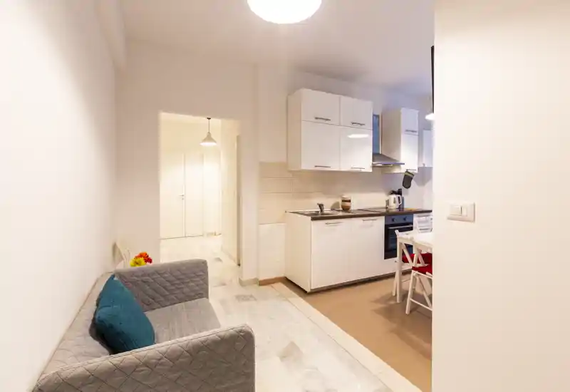 Zona giorno luminosa e compatta che si apre su una piccola cucina; divano due posti con cuscini, transizione di pavimento piastrellato e mobili cucina bianchi moderni — perfetta per un monolocale.