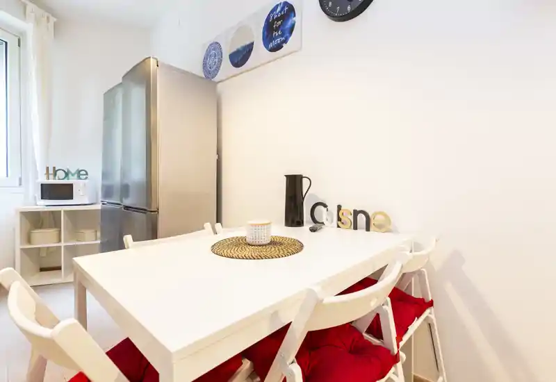 Cucina compatta e luminosa con tavolo bianco, quattro sedie con cuscini rossi, frigorifero in acciaio e microonde — perfetta per un appartamento compatto.